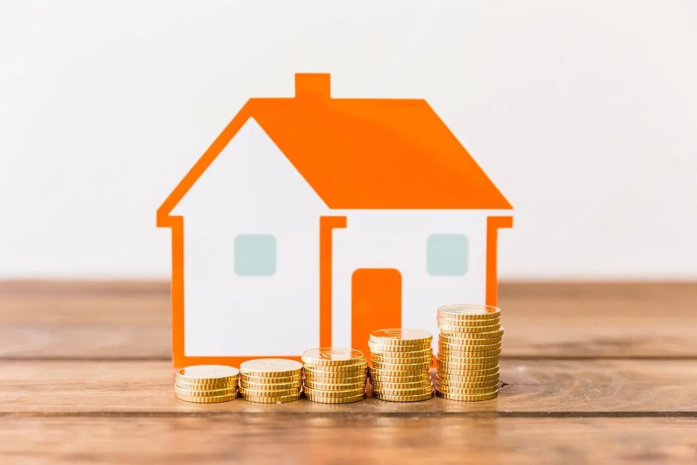 Close-up of ascending stacks of coins and a house model on a wooden desk.